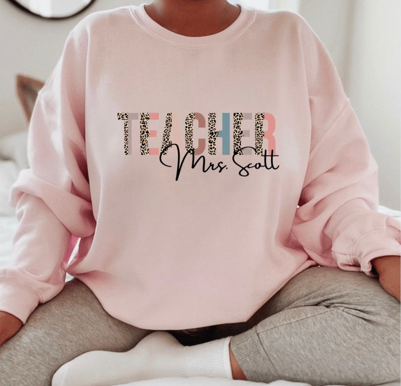 Pode incluir: Camisola cor-de-rosa com a palavra "TEACHER" em estampado de leopardo e o texto "Mrs. Scott" em it&aacute;lico.