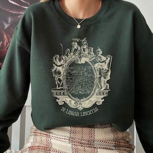 May include: A dark green sweatshirt with a vintage-style graphic print of a crest featuring figures and the text "In Libras Libertas".