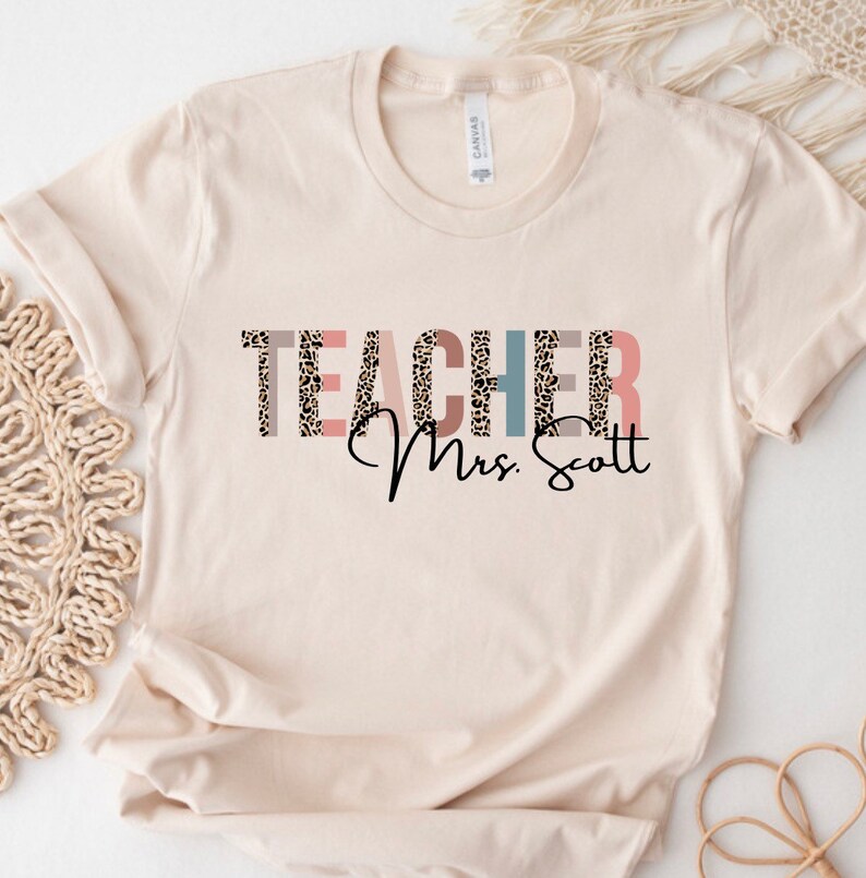 Pode incluir: Uma camiseta bege com a palavra "TEACHER" em letras de estampa de leopardo com detalhes rosa, azul e marrom. O texto "Mrs. Scott" est&aacute; escrito em it&aacute;lico preto abaixo da palavra "TEACHER".