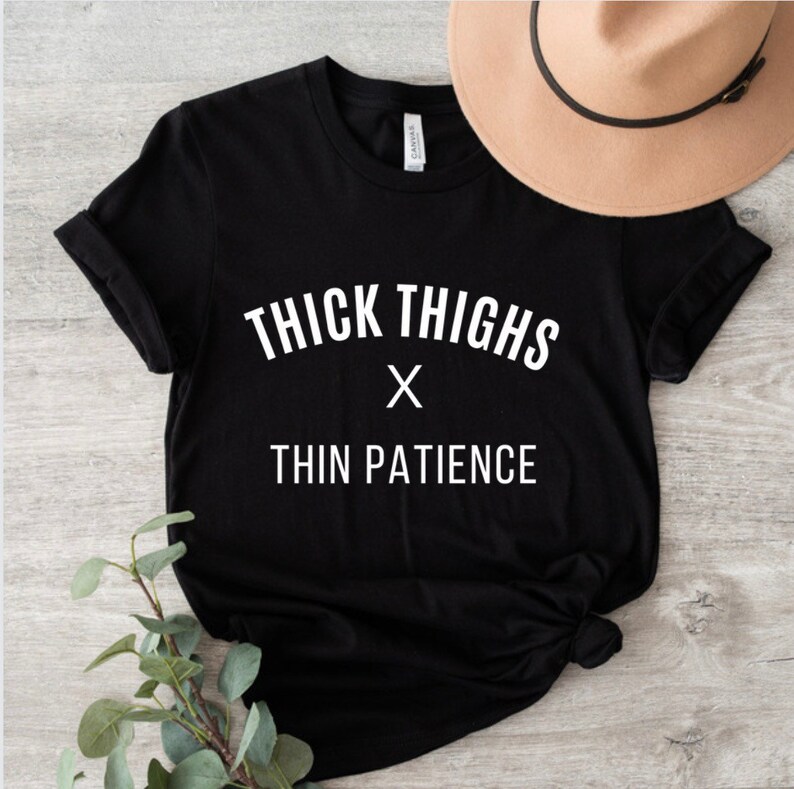 May include: Black t-shirt with white text that reads "Thick Thighs x Thin Patience".