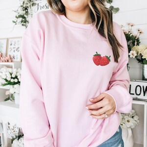 Peut inclure: Un sweat-shirt rose avec un petit motif brod&eacute; de deux fraises rouges sur la poitrine gauche.