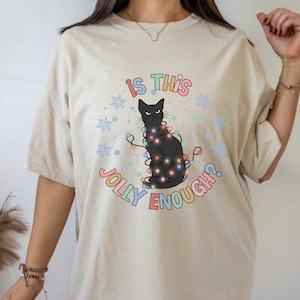 May include: A beige t-shirt with a black cat wearing Christmas lights and the text "is this jolly enough?" printed on it.