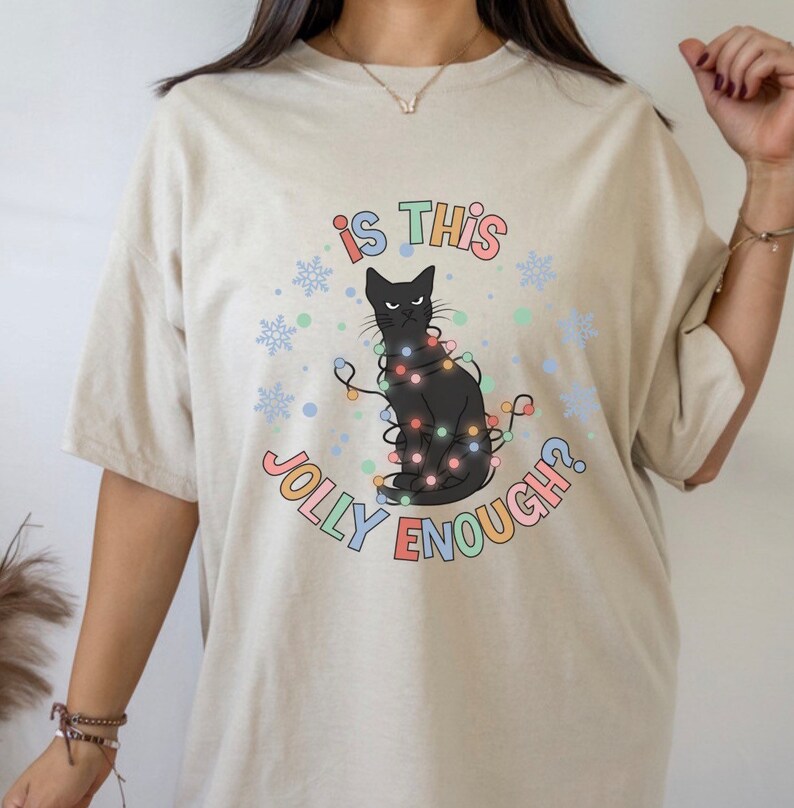 May include: A beige t-shirt with a black cat wearing Christmas lights and the text "is this jolly enough?" printed on it.