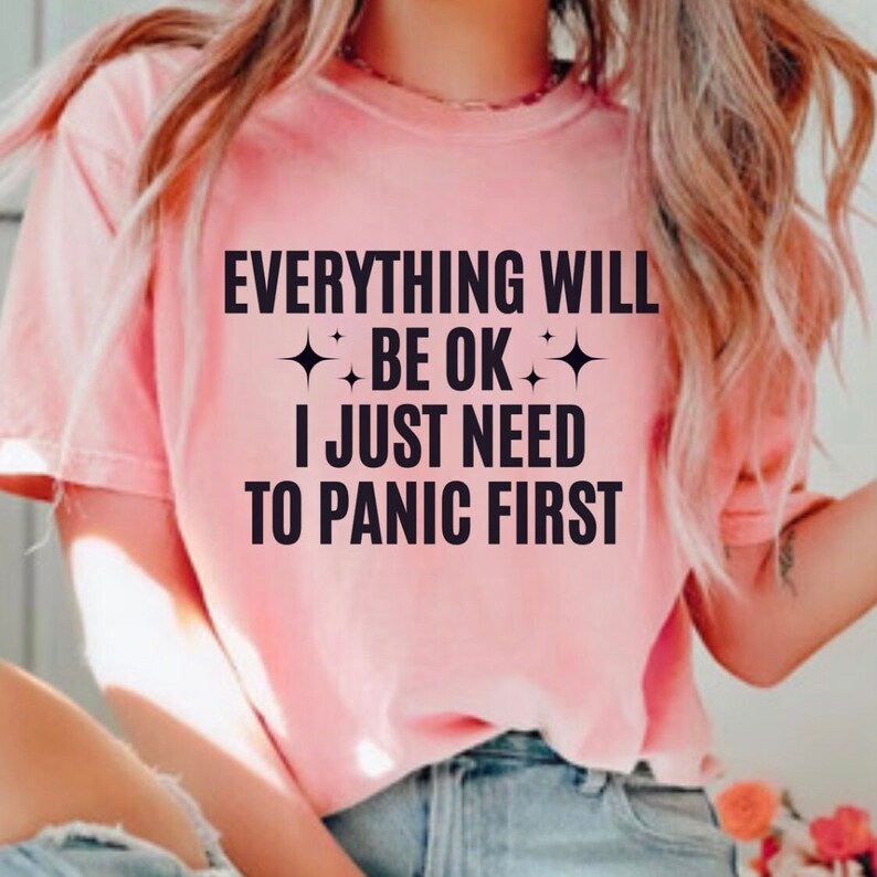 May include: A light pink t-shirt with the text "EVERYTHING WILL BE OK I JUST NEED TO PANIC FIRST" in black. The shirt has a crew neck and short sleeves.