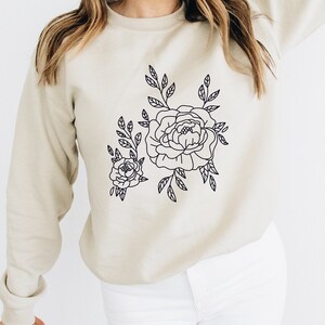 K&ouml;nnte beinhalten: Ein beiger Pullover mit einer schwarzen Umrandung eines Blumendesigns mit zwei Pfingstrosen und Bl&auml;ttern.