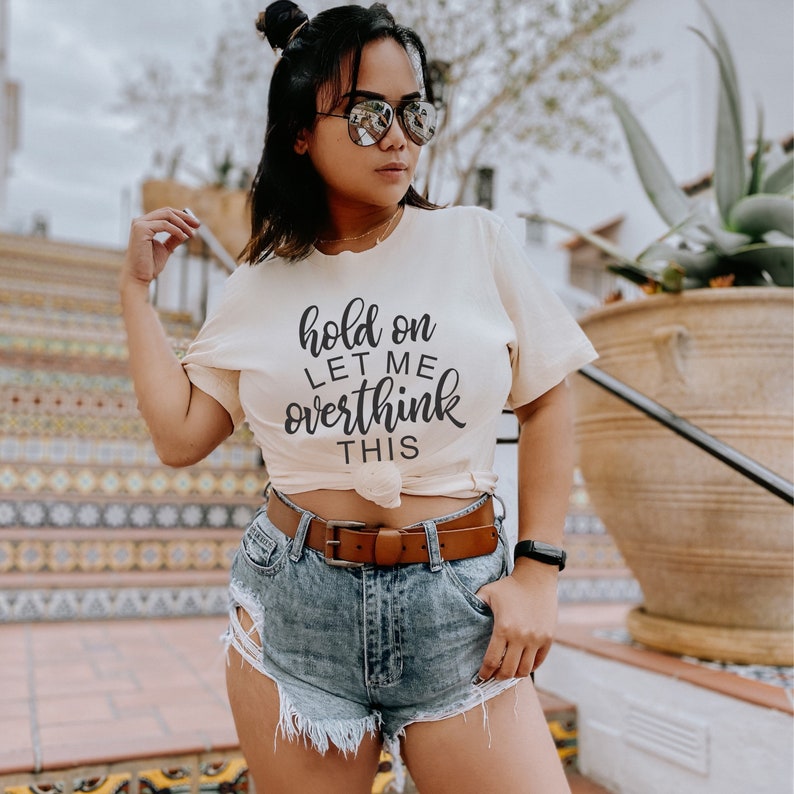 May include: A white t-shirt with the text "hold on let me overthink this" in black lettering. The shirt is being worn by a person with dark hair and sunglasses. The person is wearing denim shorts and a brown belt.