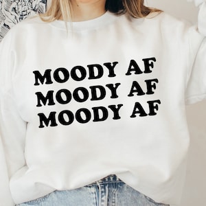 May include: White sweatshirt with the phrase "MOODY AF" repeated three times in black bold letters.