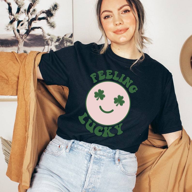 Puede incluir: Una camiseta negra con un gr&aacute;fico de cara sonriente rosa y el texto "Feeling Lucky" en verde. La cara sonriente tiene ojos de tr&eacute;bol.