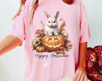 Könnte beinhalten: Ein pinkfarbenes T-Shirt mit einem weißen Kaninchen, das auf einem Kürbis mit einem grinsenden Gesicht sitzt. Der Kürbis ist von Herbstblättern und kleineren Kürbissen umgeben. Der Text "Hoppy Halloween" ist unter dem Bild gedruckt.