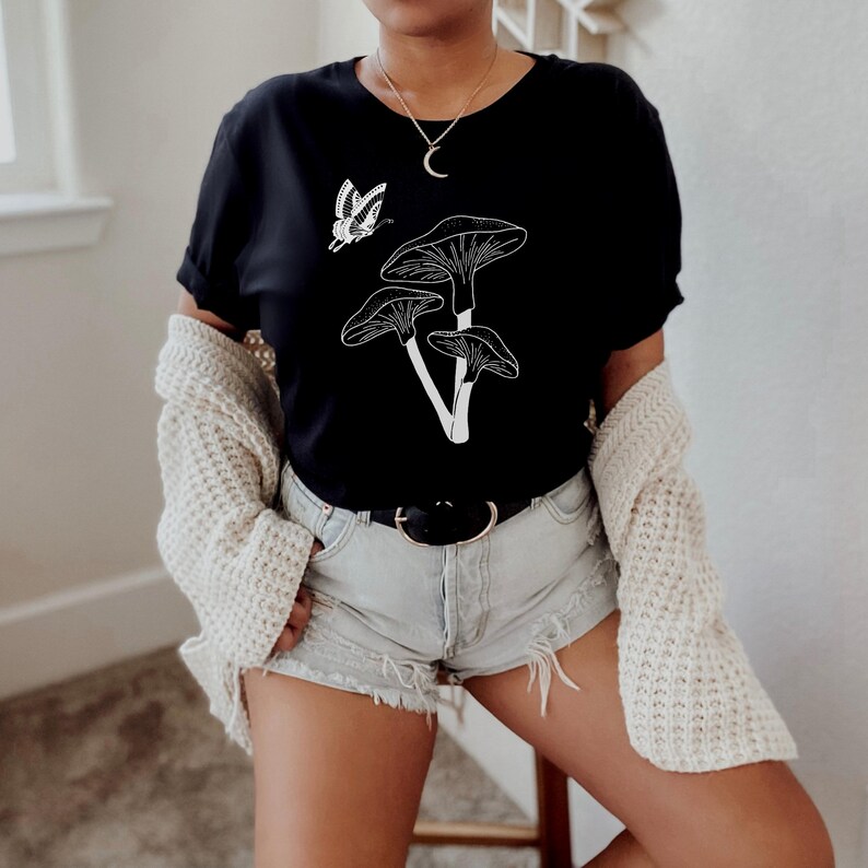May include: A black t-shirt with a white graphic of mushrooms and a butterfly. The shirt has a crew neck and short sleeves.