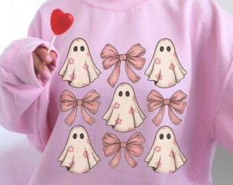 May include: A light pink sweatshirt featuring a pattern of ghost illustrations with small pink flowers and pink bows. The ghosts have two black dot eyes. The sweatshirt is a soft, comfortable material.
