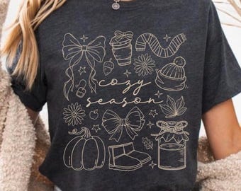Könnte beinhalten: Dunkelgraues T-Shirt mit beigem Grafikdesign mit herbstlichen Illustrationen. Das Design umfasst einen Kürbis, Schleifen, eine Kaffeetasse, einen Schal, eine Mütze und die Worte "cozy season."