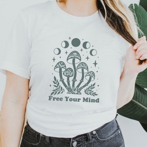 以下が含まれることがあります： 「Free Your Mind」というテキストと、キノコと月のグラフィックが描かれた白いTシャツ。