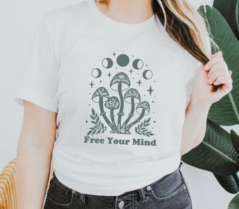 以下が含まれることがあります： 「Free Your Mind」というテキストと、キノコと月のグラフィックが描かれた白いTシャツ。