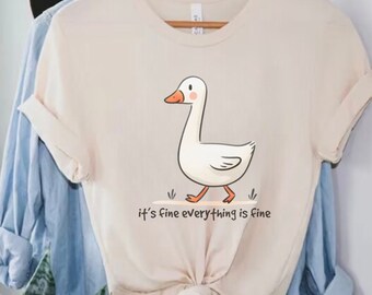 May include: A beige t-shirt featuring a cartoon white goose walking on a green patch of grass. The text "it's fine everything is fine" is printed below the goose.