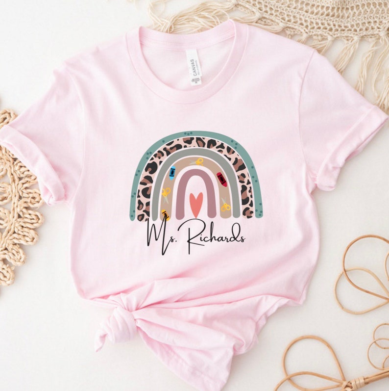 May include: A light pink t-shirt featuring a rainbow design with a leopard print pattern and school supplies. The text "Ms. Richards" is printed below the rainbow. The shirt is made of a soft material.
