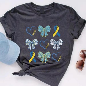 Op de afbeelding: Een donkergrijs T-shirt met een patroon van blauwe en gele linten, harten en stippen. De linten zijn gebonden in strikken.