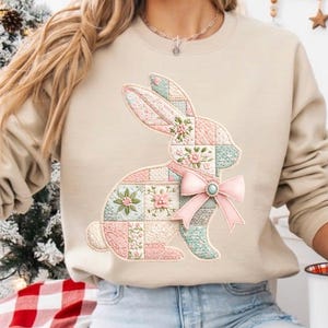 Può includere: Felpa beige con un design a coniglietto patchwork nei toni pastello rosa, blu e bianco. Il coniglietto ha un fiocco rosa con un centro turchese. Il design include dettagli floreali e trapuntati, perfetto per Pasqua o la primavera.