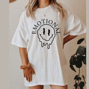 May include: White oversized t-shirt with a black dripping smiley face graphic and the text "EMOTIONAL" around it.