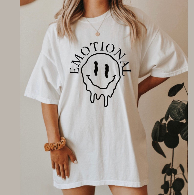 May include: White oversized t-shirt with a black dripping smiley face graphic and the text "EMOTIONAL" around it.