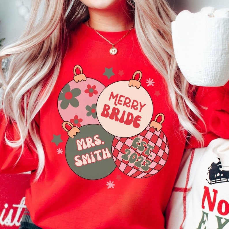 K&ouml;nnte beinhalten: Roter Weihnachtspullover mit drei Christbaumkugeln. Die Kugeln sind mit rosa, gr&uuml;nen und roten Karomustern verziert. Der Text auf den Kugeln lautet "Merry Bride", "Mrs. Smith" und "Est. 2023".