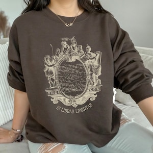 May include: A brown sweatshirt featuring a vintage-style graphic print of a crest with the text "In Libras Libertas".
