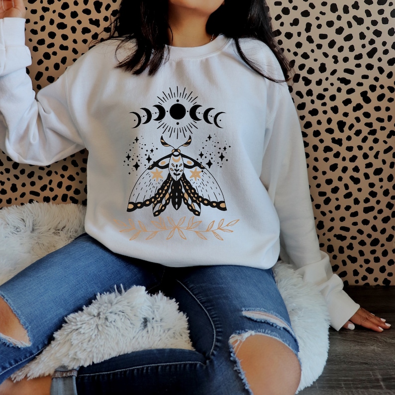May include: White sweatshirt with a black and gold moth design, moon phases, and stars. The moth has a floral pattern on its wings.