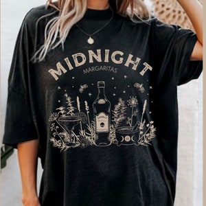 May include: Black t-shirt with the text "MIDNIGHT MARGARITAS" in white. The shirt features a graphic of a bottle of tequila, a margarita glass, and a mortar and pestle with herbs and flowers.