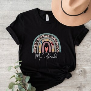 May include: Black t-shirt featuring a colorful rainbow design with a leopard print pattern and school-themed icons. The name "Ms. Richards" is written in cursive below the rainbow. A tan hat is visible in the background.