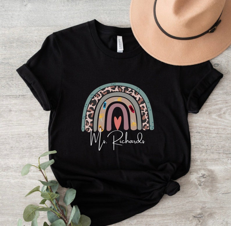 May include: Black t-shirt featuring a colorful rainbow design with a leopard print pattern and school-themed icons. The name "Ms. Richards" is written in cursive below the rainbow. A tan hat is visible in the background.