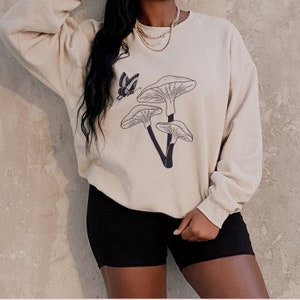 May include: A beige sweatshirt with a graphic of mushrooms and a butterfly. The sweatshirt has long sleeves and a crew neck.