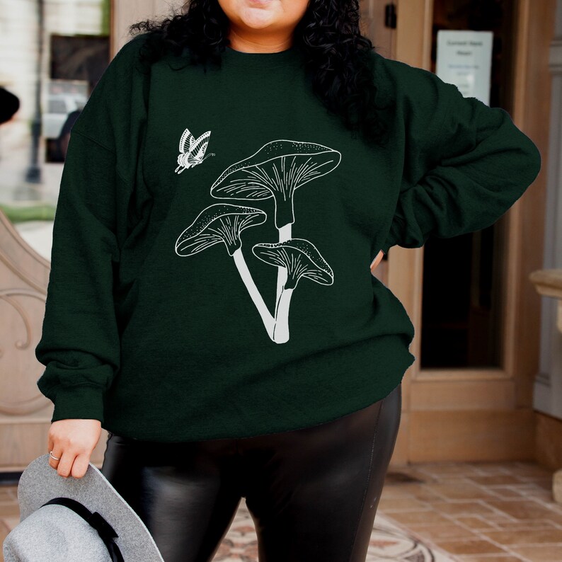 May include: A dark green sweatshirt with a white outline of mushrooms and a butterfly. The sweatshirt has a crew neck and long sleeves.