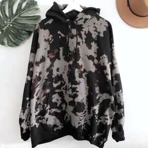 May include: A long-sleeve hooded sweatshirt featuring a black and grey tie-dye pattern. The garment includes a front kangaroo pocket, a drawstring hood with metal eyelets, and ribbed cuffs at the sleeves and hem, displayed on a white hanger.