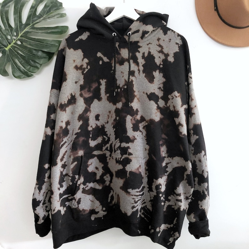 May include: A long-sleeve hooded sweatshirt featuring a black and grey tie-dye pattern. The garment includes a front kangaroo pocket, a drawstring hood with metal eyelets, and ribbed cuffs at the sleeves and hem, displayed on a white hanger.