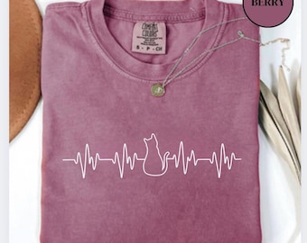 Può includere: Una maglietta color bacca con un contorno bianco di un gatto seduto su una linea di battito cardiaco. La maglietta ha un piccolo cartellino che dice "Comfy Colors" e "S - P - Ch".