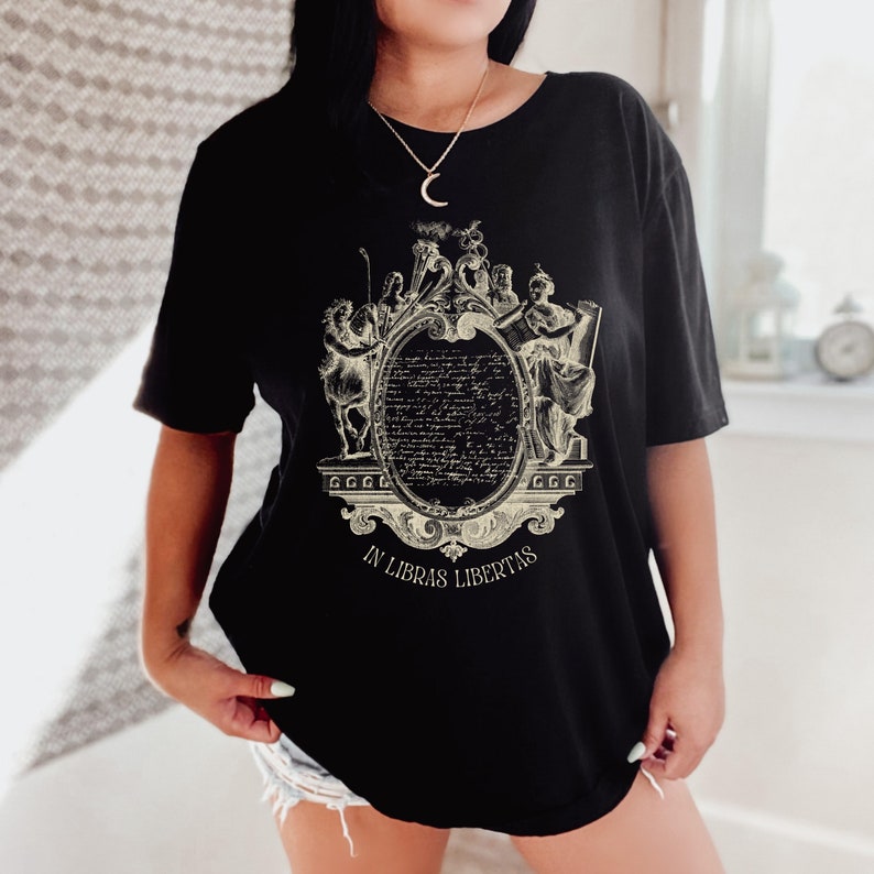 May include: Black t-shirt with a cream-coloured graphic of an ornate frame and text. The frame surrounds a text-filled oval. The words "IN LIBRAS LIBERTAS" are printed below the frame. The shirt is a casual style.