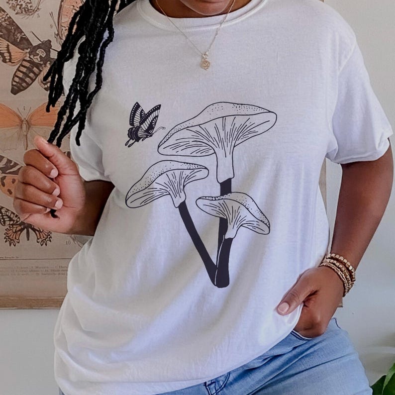 May include: White t-shirt with a black line drawing of three mushrooms and a butterfly.