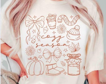 Könnte beinhalten: Cremefarbenes T-Shirt mit braunem Grafikdesign mit herbstlichen Illustrationen. Das Design umfasst Schleifen, eine Kaffeetasse, einen Schal, Blumen, eine Mütze, einen Kürbis, Stiefel und die Worte "cozy season."