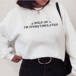 Może przedstawiać: Biała bluza z czarnym napisem "HOLD ON I'M OVERSTIMULATED" z małymi gwiazdkami po obu stronach tekstu.
