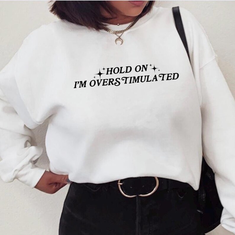 Może przedstawiać: Biała bluza z czarnym napisem "HOLD ON I'M OVERSTIMULATED" z małymi gwiazdkami po obu stronach tekstu.