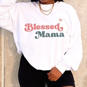 Puede incluir: Sudadera blanca con un gr&aacute;fico de fuente retro rosa y turquesa que dice "Blessed Mama" con un peque&ntilde;o coraz&oacute;n rosa sobre el texto.