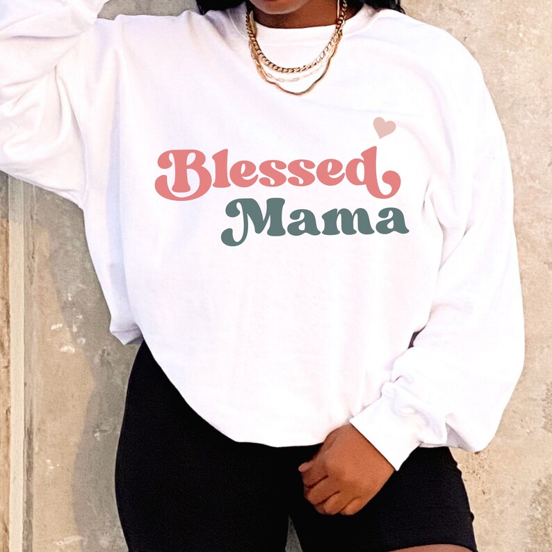 Puede incluir: Sudadera blanca con un gr&aacute;fico de fuente retro rosa y turquesa que dice "Blessed Mama" con un peque&ntilde;o coraz&oacute;n rosa sobre el texto.