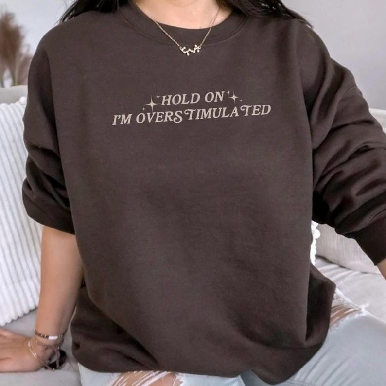 Może przedstawiać: Brązowa bluza z napisem "HOLD ON I'M OVERSTIMULATED" w białym kolorze.