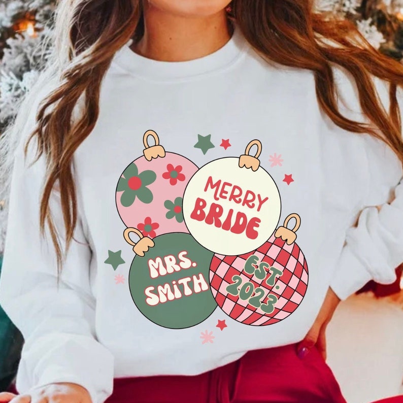 K&ouml;nnte beinhalten: Wei&szlig;er Pullover mit einem Weihnachtsbaumschmuck-Design. Der Baumschmuck ist rosa, gr&uuml;n und rot mit wei&szlig;en Punkten. Der Text auf dem Baumschmuck lautet "Merry Bride", "Mrs. Smith" und "Est. 2023".