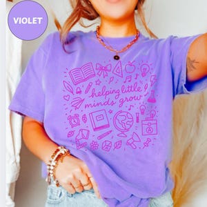 Puede incluir: Una camiseta violeta con un gráfico rosa que dice "helping little minds grow". El diseño incluye iconos relacionados con la escuela, como libros, un globo terráqueo y una bombilla. La camiseta tiene mangas cortas y cuello redondo.