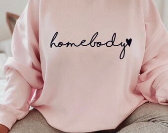 Puede incluir: Sudadera rosa claro con la palabra "homebody" en fuente negra y un pequeño corazón negro. La sudadera es de cuello redondo y parece estar hecha de un material suave.