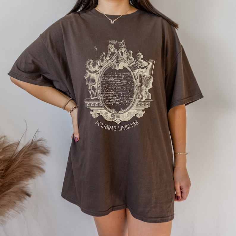May include: An oversized brown t-shirt with a vintage-style graphic print of a crest featuring a scroll with the text "In Libras Libertas".