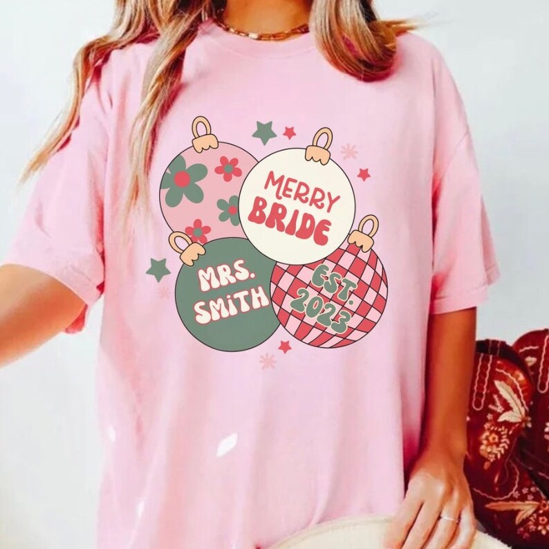 K&ouml;nnte beinhalten: Pinkes T-Shirt mit einem Motiv aus drei Weihnachtskugeln. Die Kugeln sind mit gr&uuml;nen, roten und wei&szlig;en Mustern verziert. Der Text auf den Kugeln lautet "Merry Bride", "Mrs. Smith" und "Est. 2023".