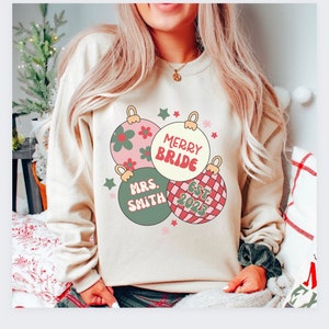 K&ouml;nnte beinhalten: Ein cremefarbener Pullover mit einem Weihnachtsbaumschmuck-Design. Der Baumschmuck ist rosa, gr&uuml;n und rot mit wei&szlig;en Akzenten. Der Text auf dem Pullover lautet "Merry Bride", "Mrs. Smith" und "Est. 2023".