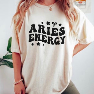May include: Cream-colored t-shirt with the words "ARIES ENERGY" in bold, black, retro-style font. The design includes star and ram symbols. The shirt is a casual, oversized fit.